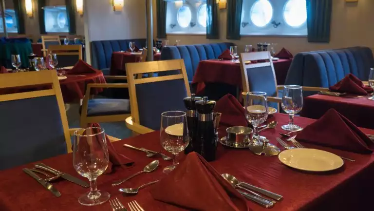 Dining room of Quest expedition ship set for dinner with burgundy tablecloths, wood chairs, blue booths & porthole windows.