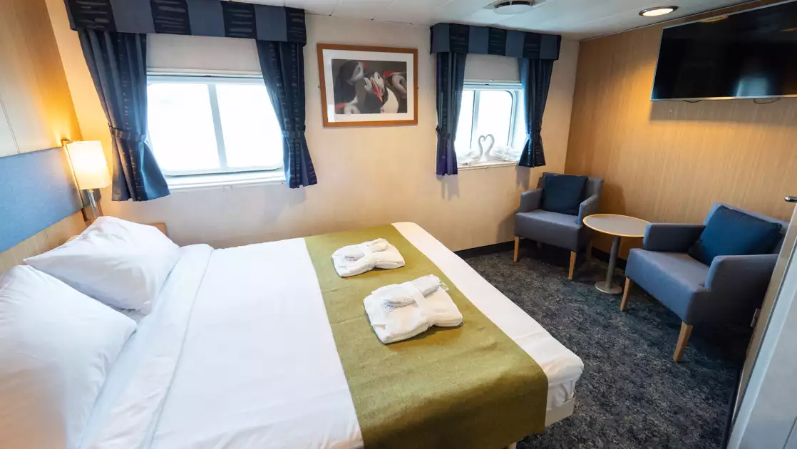 Double bed in white & green linens by 2 windows, desk & blue chairs in Double Cabin Superior aboard Quest Arctic expedition ship.