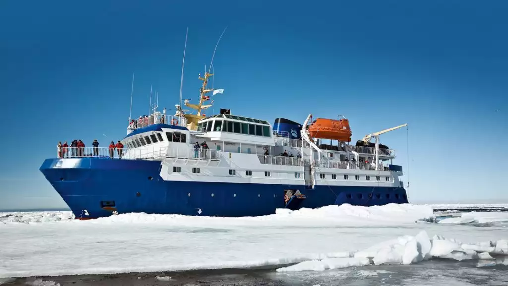 Quest arctic small ship with blue hull, white upper decks & orange life boat sits among ice on a sunny day.