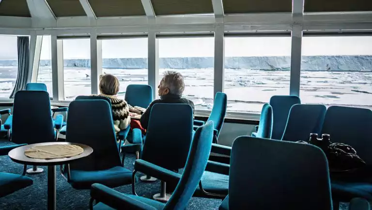 2 guests sit in lounge of Quest expedition ship with small circular coffee tables, high-back teal chairs & large view windows.