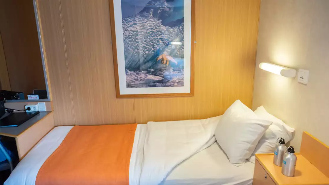 Twin bed in white & salmon-colored linens under reading light, by bedside table in Twin Cabin for single use aboard Quest ship.