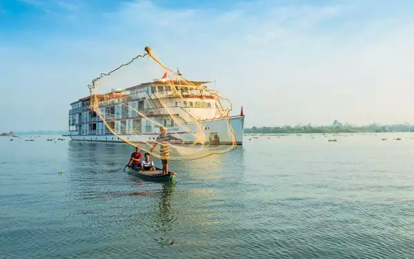 Jahan ship with white decks & red trim sits behind canoe of 3 fishermen casting a net in the sun, seen on southeast asia cruises.