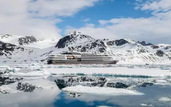 A luxury ship colored white and silver with a red stripe float in ice covered water in front of snowy mountain range in Svalbard