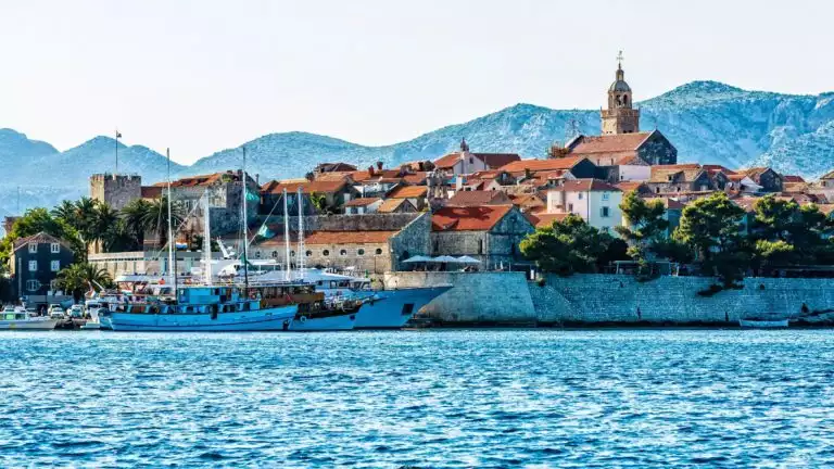 water view of a small costal town with a large sailboat parked next to the city walls and lush green trees