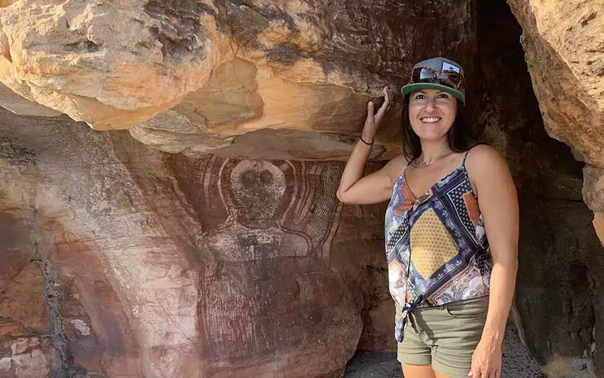 Woman in tank top, shorts & hat stands beside ancient rock paintings seen on Kimberley Australia cruises.