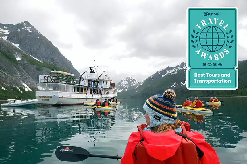 Girl wearing colorful beanie kayaking in Alaska amongst other kayakers headed towards a small white wooden ship. 