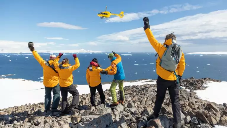 Group of guests in yellow coats stand atop rocky summit & wave to yellow helicopter on a sunny day in Antarctica.