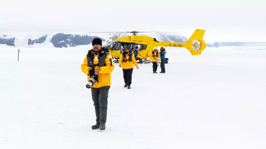 Group of guests in yellow coats on an Antarctica cruise with helicopter walk over snow beside a yellow heli during a landing.