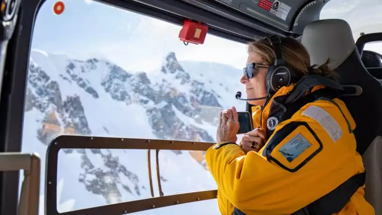 Woman in yellow jacket looks out the window to jagged, snow-covered peaks during an Antarctica helicopter tour.