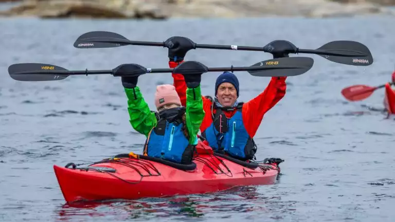 Man & girl raise kayak paddles while in a red boat during the Arctic Golden Autumn & Northern Lights trip.