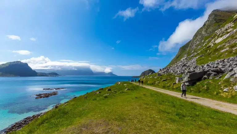 Arctic Golden Autumn & Northern Lights traveler walks a flat road by green grass & teal sea on a sunny day in Norway.