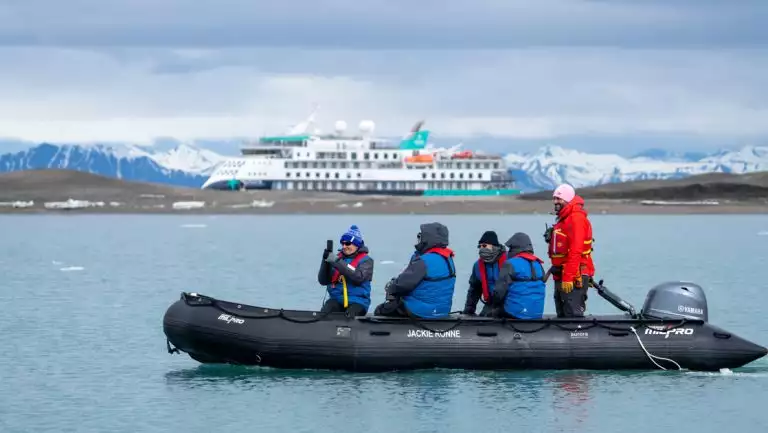 Zodiac with Arctic Golden Autumn & Northern Lights travelers moves through calm sea while guests take photos as ship sits nearby.