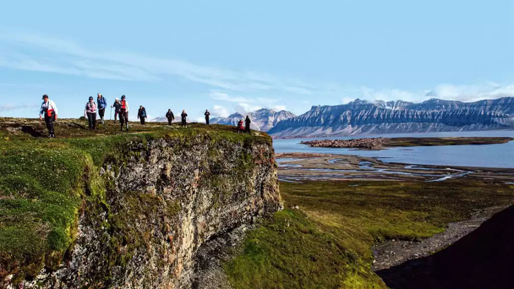 Small group of Expedition Svalbard cruise guests hike a grassy green bluff overlooking a peaceful fjord on a sunny day.