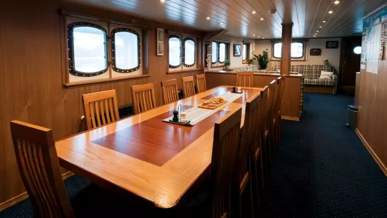 Dining room on MS Svojeien with long wood table with 12 high-back chairs atop blue carpet by oval windows & lounge behind.