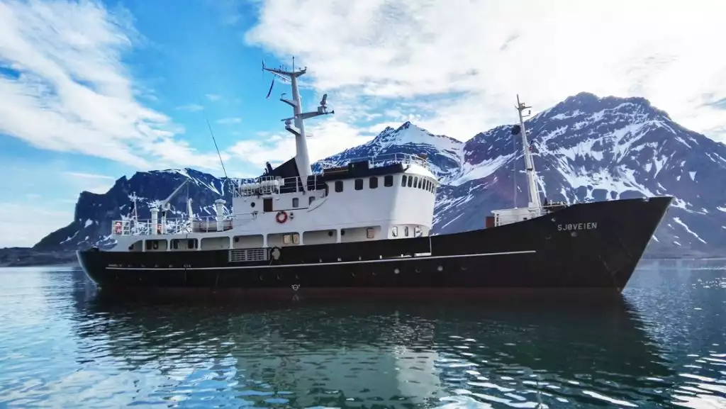 MS Sjoveien Arctic ship with black & red hull, white upper decks & 2 masts, sitting near snowy peaks in calm sea.