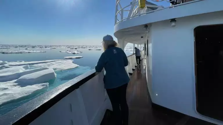 Female Svalbard traveler stands on deck of MS Svojeien with its white & black paint as it cruises past icebergs in the sun.