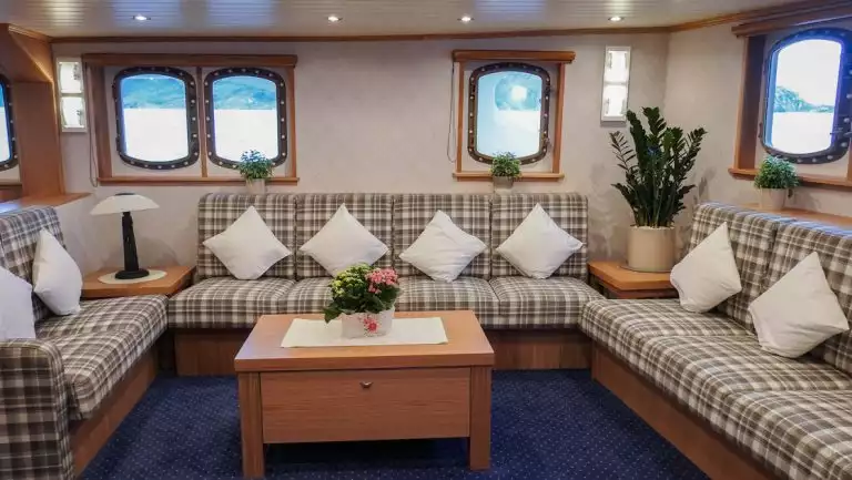 Wraparound plaid couch with white throw pillows by wood coffee table atop blue carpet under oval windows aboard MS Svojeien.