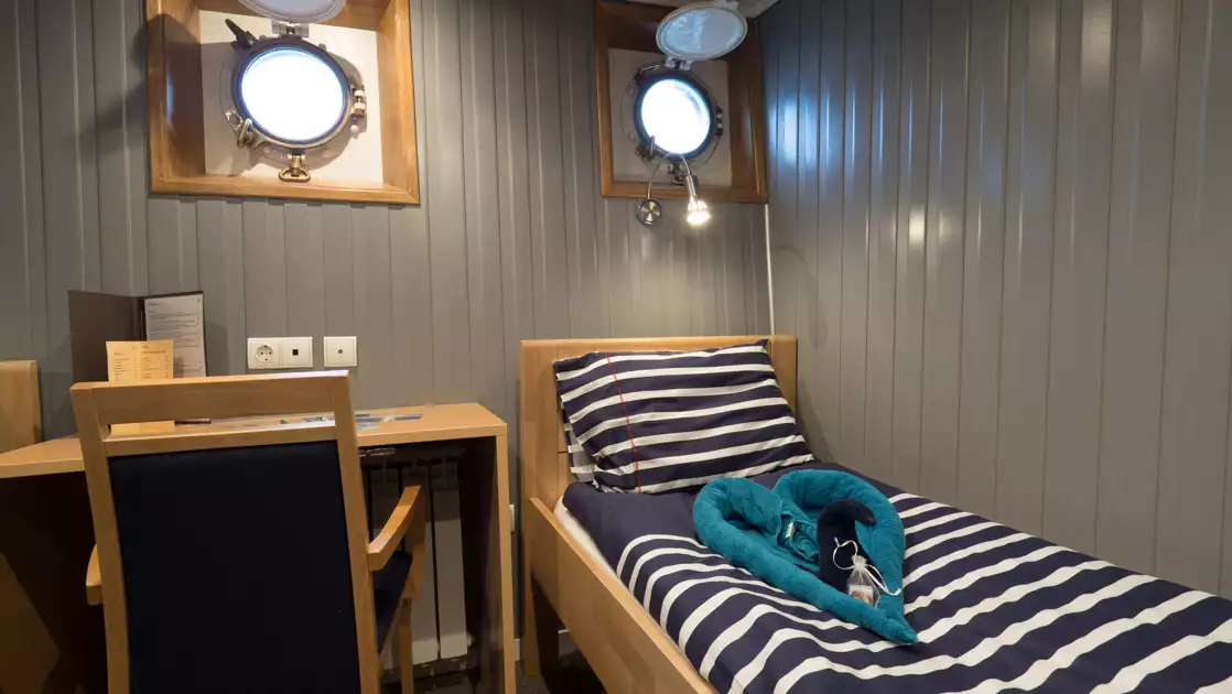 Twin cabin with 1 bed in nautical colors under reading light with small desk beside, 2 portholes & gray walls on MS Svojeien.