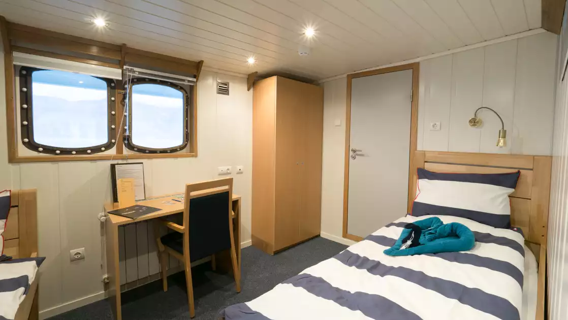 Twin cabin with 2 beds in nautical colors under reading lights, 2 windows, small desk, armoire & white walls on MS Svojeien.