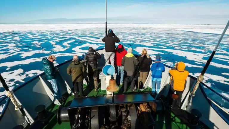 Small group of Svalbard travelers in bright coats of varying colors stand on bow of MS Stockholm ship & cruise through ice.