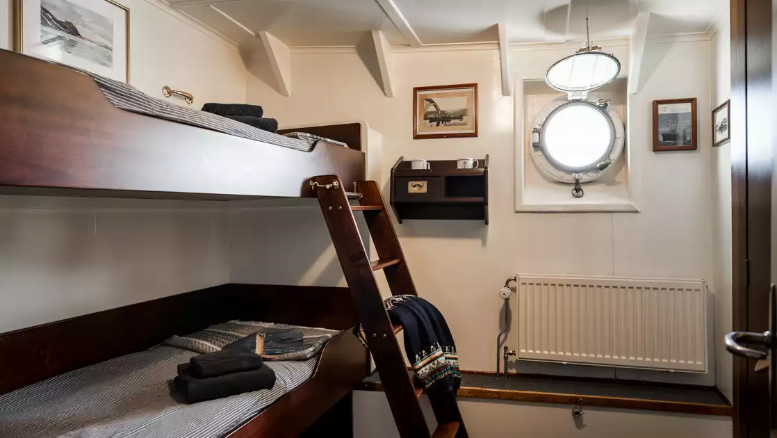 Cabin 25 on MS Stockholm ship with wood bunk beds, porthole, white walls & radiator, framed old photos & door to bathroom.