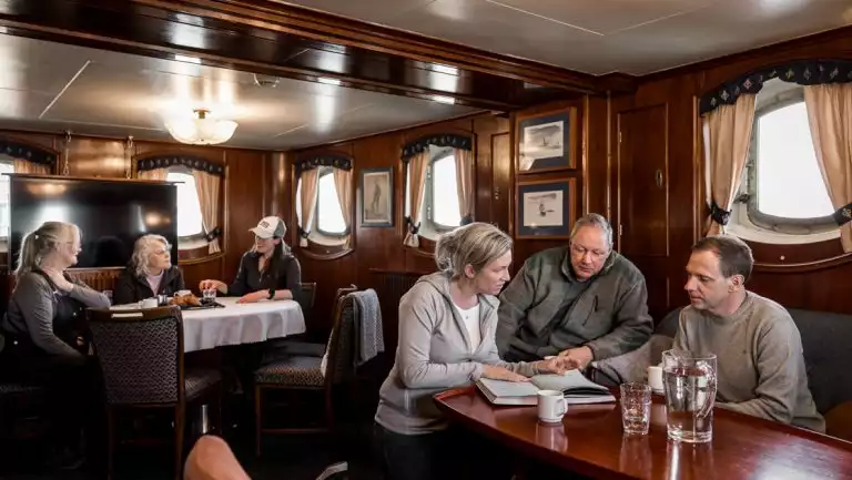 5 guests & 1 guide sit at tables in wood-paneled lounge with view windows & classic decor on MS Stockholm ship.