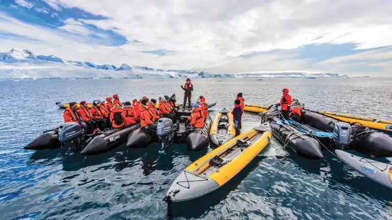 Travelers in orange jackets prepare to venture out in small skiffs and kayaks on a sunny and cloudy day