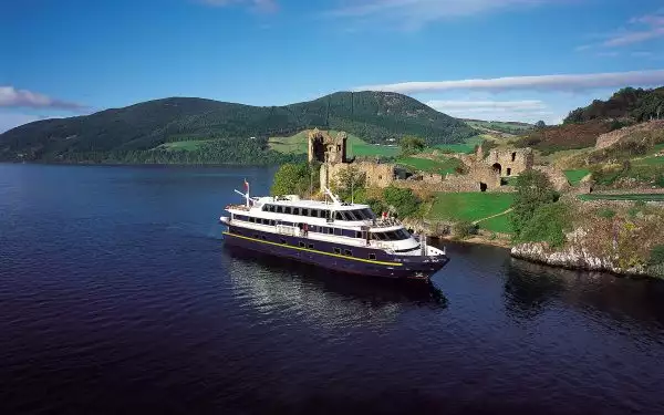 An small Northern Europe river cruise ship with a blue hull sails along an isolated Scotland coastline with a castle on it