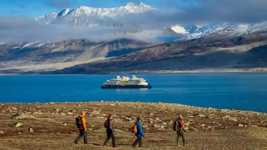 Four hikers on a Northern Europe tour in Greenland hike on a brown seaside slope with a small ship in the background