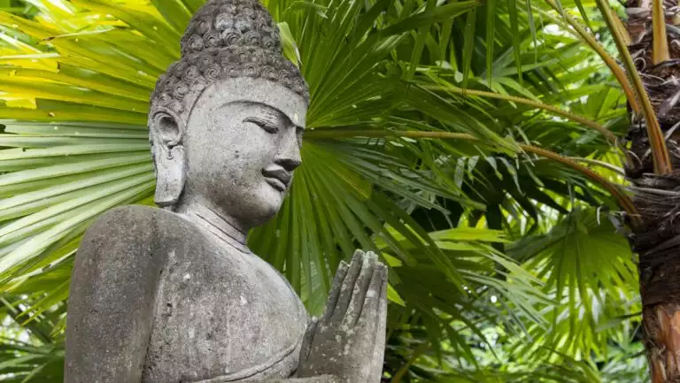 Weather gray stone Buddha statue with green palms behind seen on a Paspaley Pearl East Indonesia cruise.