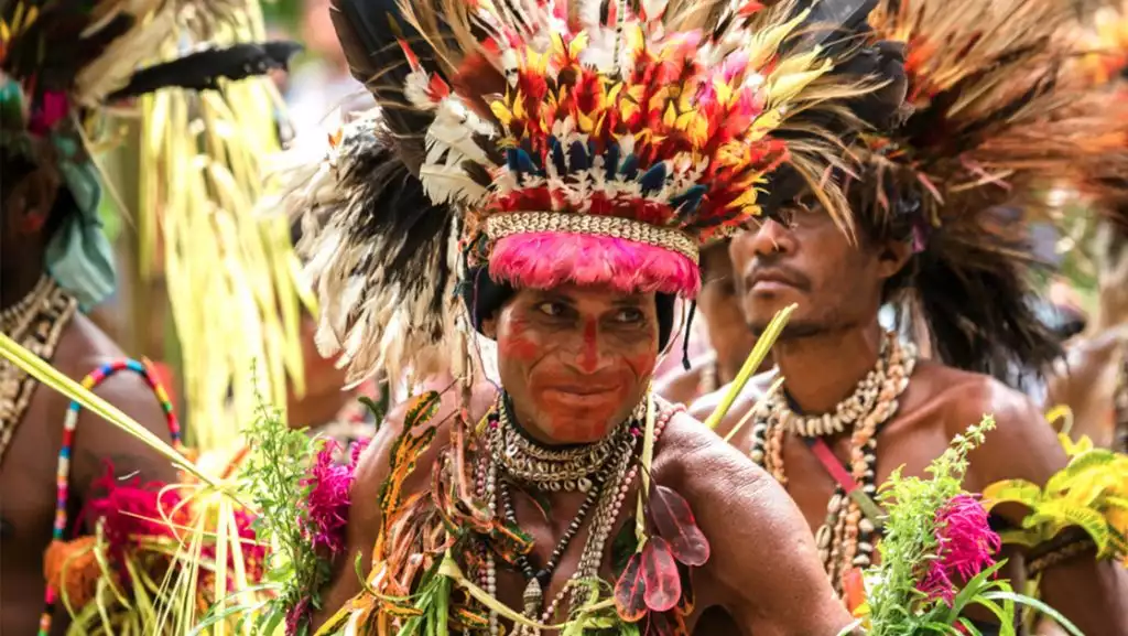 Indigenous man in face paint, ceremonial feather headdress, shell necklaces & leaf garb looks across the dance troupe.