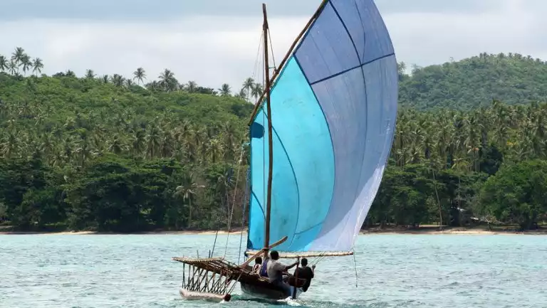 Locals in Papua New Guinea sail and outrigger canoe in teal sea near a shoreline of lush green jungle on a sunny day.