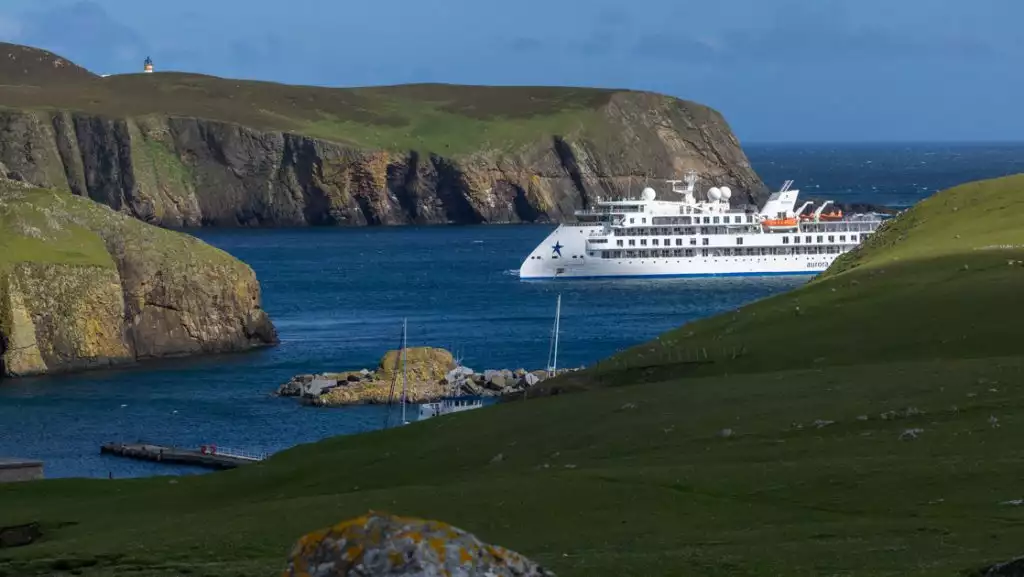 Modern, white small ship sits in inlet by grassy rolling hills with rocky cliffs on a Scotland, Faroes & Iceland cruise.