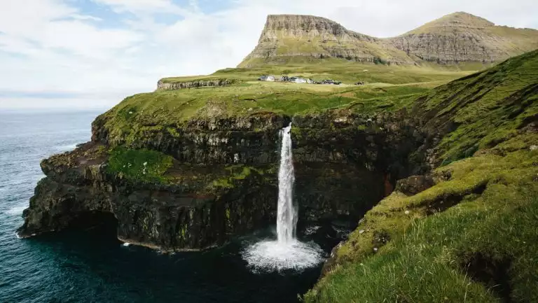 Mulafossur waterfall cascades from grassy knoll over rock cliffs to sea below, as mountains rise behind in the Faroe Islands.