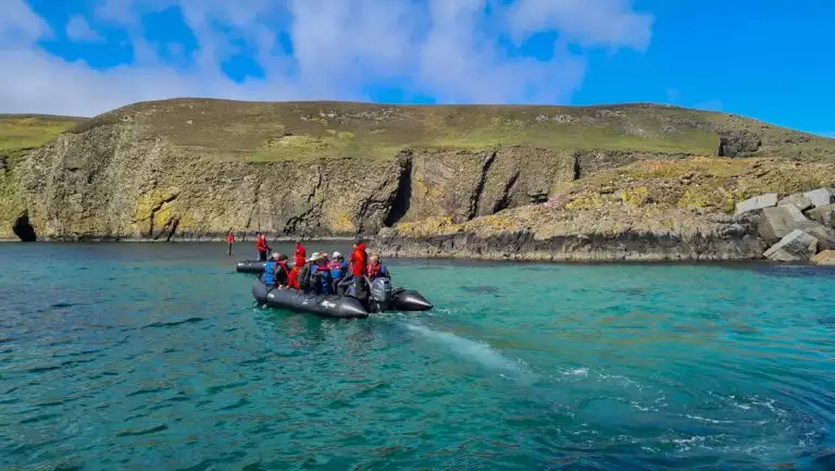 2 Zodiac boats with Scotland, Faroes & Iceland travelers in red & blue coats motor toward rocky shoreline with grass & flowers.