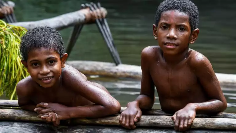 2 young boys stand in water & hold onto a primitive outrigger canoe during a Sepik River cruise in Papua New Guinea.