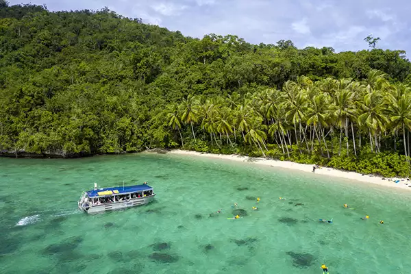 A small ship sails towards a tropical shoreline in Papua New Guinea with snorkelers in the clear green waters and lush palm trees on the shore