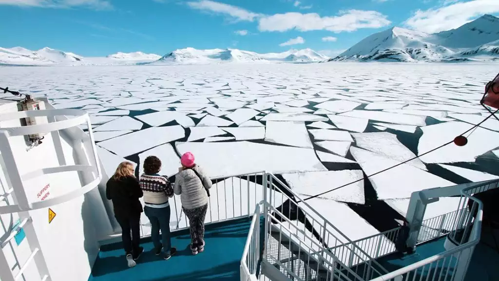 3 Arctic travelers stand on deck of small ship & view large chunks of ice with snowy peaks behind on a sunny day in Svalbard.