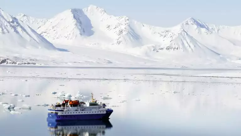 Quest polar expedition ship with blue hull & white upper decks sits in glassy sea by snow-covered mountains in Svalbard.