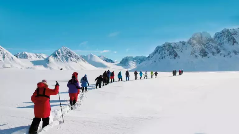 Small group of Svalbard travelers walk a single-file line using snowshoes near snowy peaks on a sunny day in the Arctic.