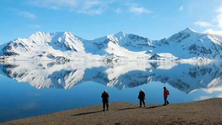 3 Svalbard Adventure Cruise guests walk a rocky overlook above glassy sea & snow-capped mountains in full arctic sunshine.