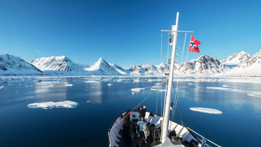 A few Svalbard Adventure Cruise guests stand on bow of small white ship, viewing snowy peaks, icebergs & glassy sea in the sun.