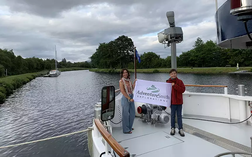 A female traveler stands with her young son on the bow of a small ship on a Scotland canal holding a white AdventureSmith Explorations flag