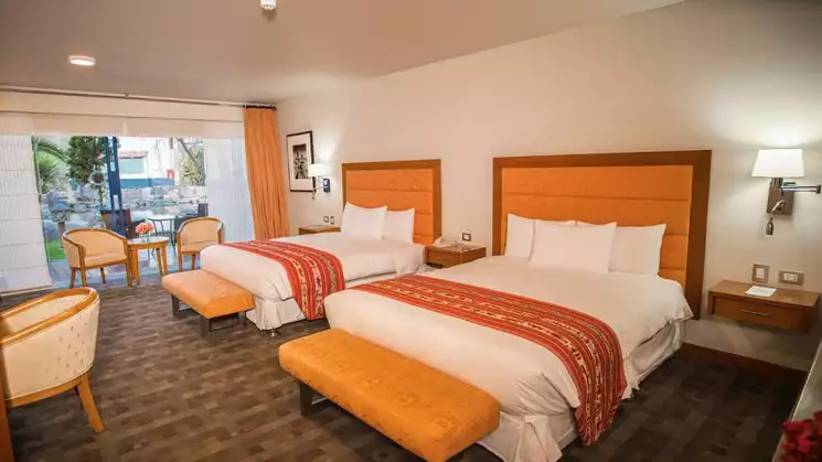 Standard hotel bedroom with two tan beds with white sheets and white walls with brown carpet and side tables and chairs