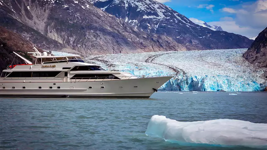 A small luxury Alaska cruise ship is seen at anchor in front of a big tidewater glacier with an iceberg in the water in front of the yacht