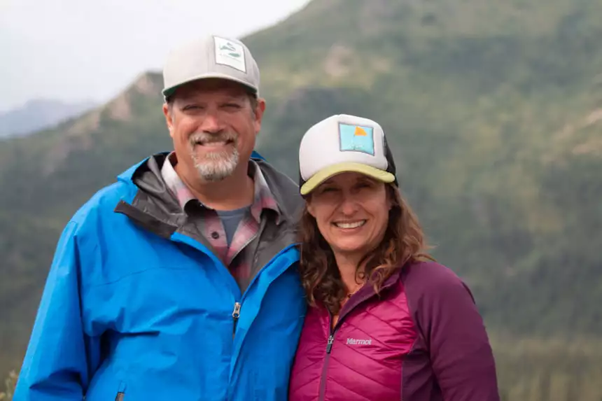 Man in blue jacket & gray hat stands with woman in berry-colored coat & light cap in Denali, Alaska.