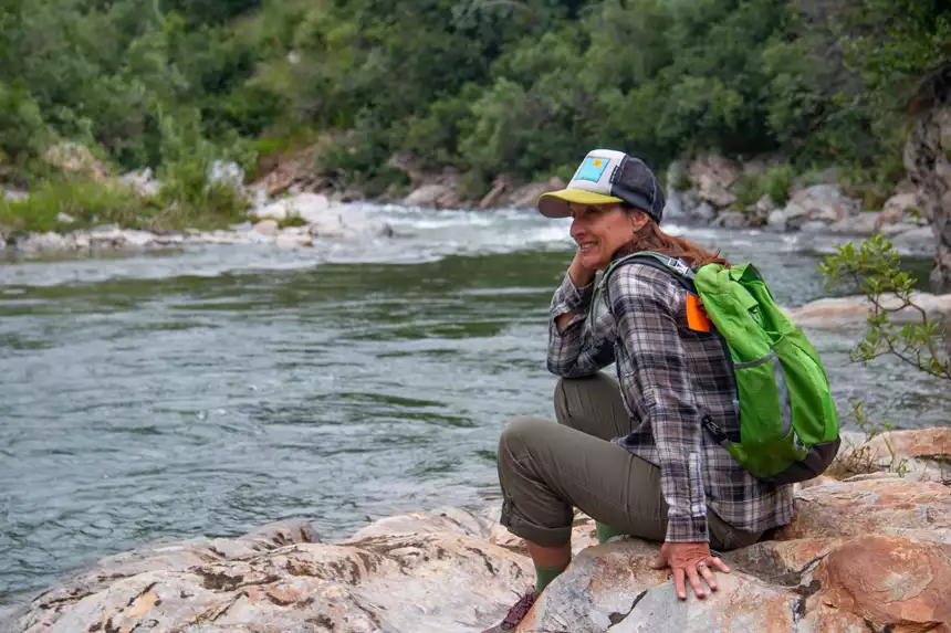 Woman in plaid shirt & hiking gear sits on rocks beside a gentle river during a visit to Denali National Park.