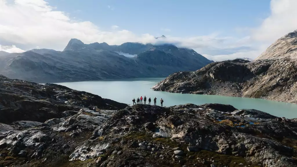 Aerial view of small group of hikers standing atop a rocky mountain overlooking a fjord with teal water in East Greenland.