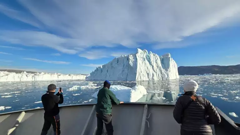 3 East Greenland Sermilik fjord cruise guests stand on bow of small ship in warm clothes, photographing large icebergs.