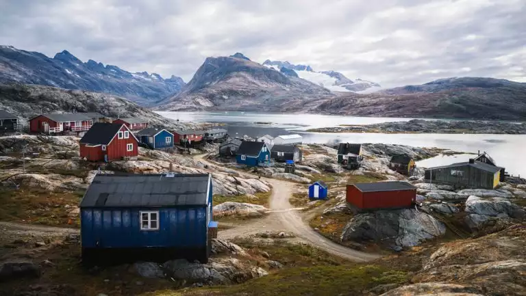 Small coastal town with colorful buildings, green tundra & bare rock under cloudy skies in southeast Greenland.